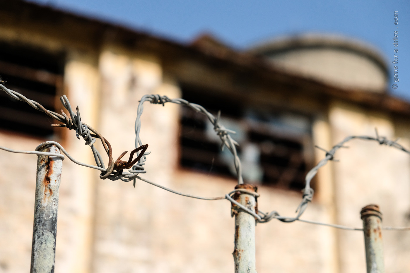 Nahaufnahme eines Stacheldrahtzaunes in Kucova, Albanien. Eine verfallene Fabrikanlage unscharf dahinter.